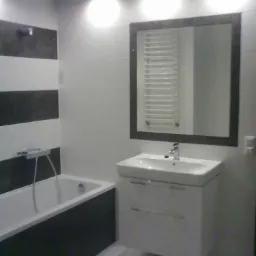 Wykończona łazienka z wanną obudowaną ciemnymi panelami, umywalką z szafką wiszącą, lustrem w ciemnej ramie i białymi płytkami z poziomymi pasami ciemnych płytek.