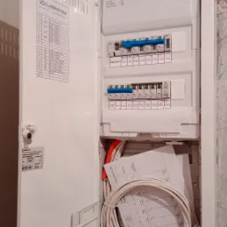 Otwarta skrzynka bezpiecznikowa z widocznymi bezpiecznikami, przewodami i schematami elektrycznymi wewnątrz, zamontowana na surowej ścianie.