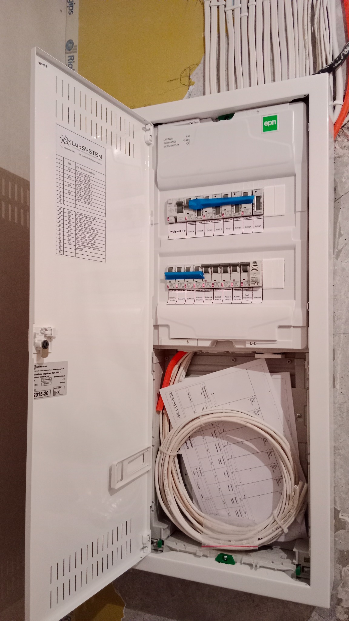 Otwarta skrzynka bezpiecznikowa z widocznymi bezpiecznikami, przewodami i schematami elektrycznymi wewnątrz, zamontowana na surowej ścianie.