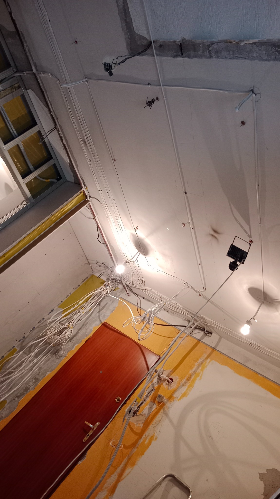 Wnętrze w trakcie remontu, widoczne liczne przewody elektryczne poprowadzone po suficie i ścianach, surowy tynk, prowizoryczne oświetlenie halogenowe, częściowo pomalowana ściana na żółto, brązowe...