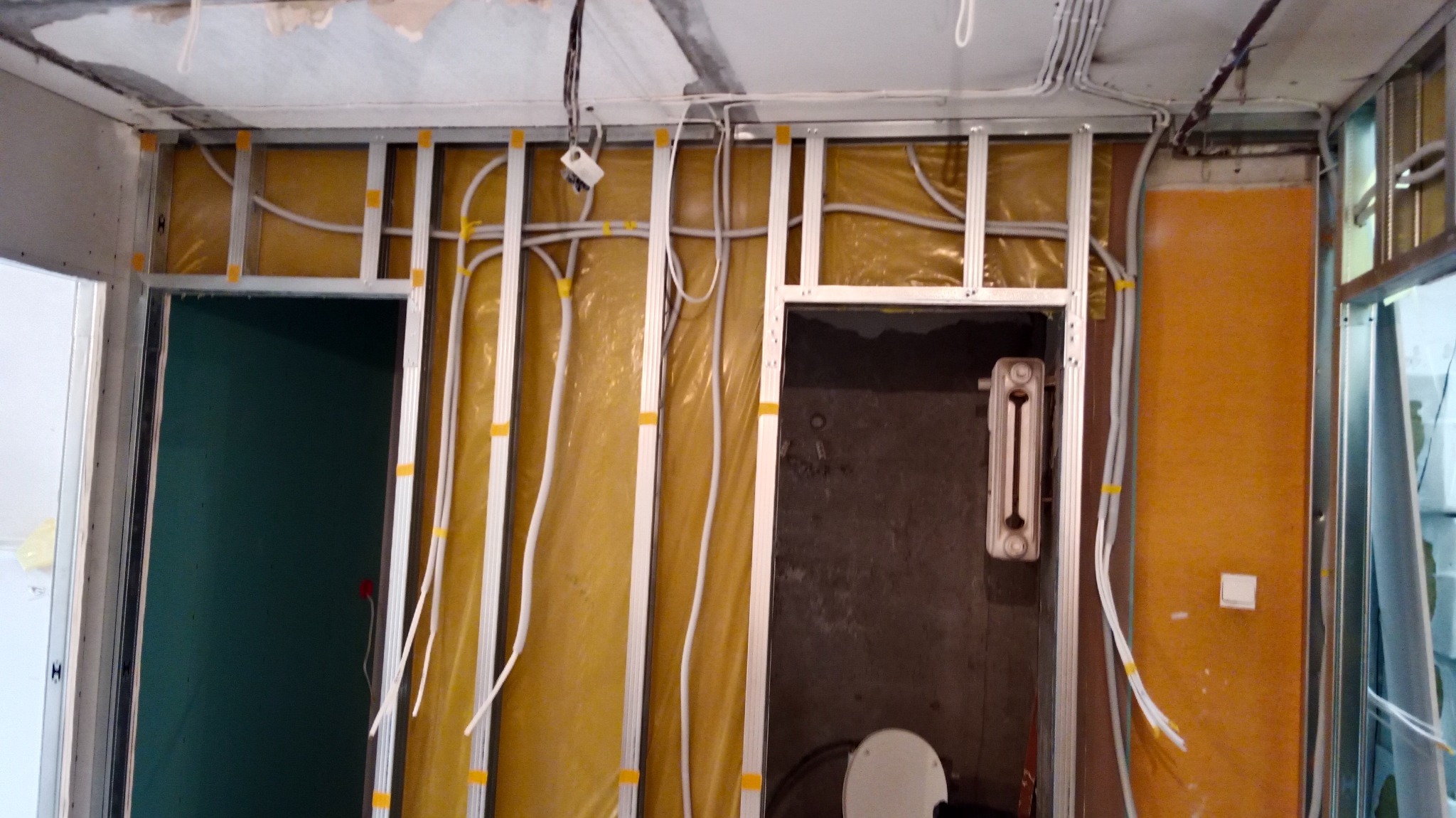 Wnętrze w trakcie remontu: widoczne profile aluminiowe, izolacja w kolorze żółtym, białe rury instalacyjne przymocowane taśmą, surowa ściana z grzejnikiem i toaletą w tle.