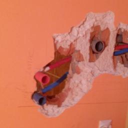 MichaelAngelo Interiors With Passion ANDRZEJ NOWAK - Odsłonięte rury instalacji wodnej (czerwona i niebieska) w wykuciu w pomarańczowej ścianie, widoczne ślady po wierceniu i resztki gruzu, fragment drewnianej podłogi po prawej stronie.