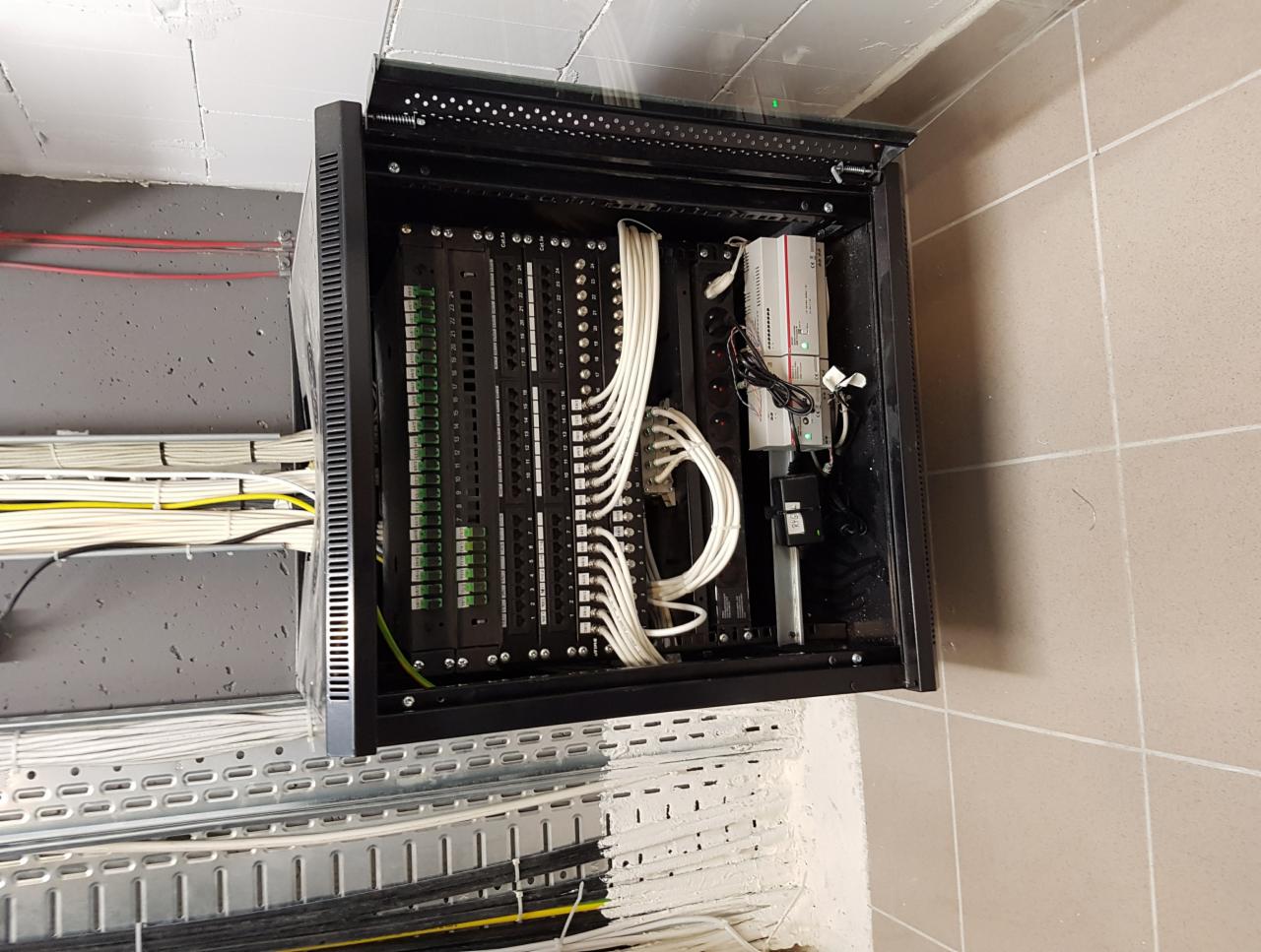 Otwarta szafa rack z panelami krosowymi i okablowaniem strukturalnym, zamontowana na ścianie w pomieszczeniu z widocznymi kablami w korytach kablowych.