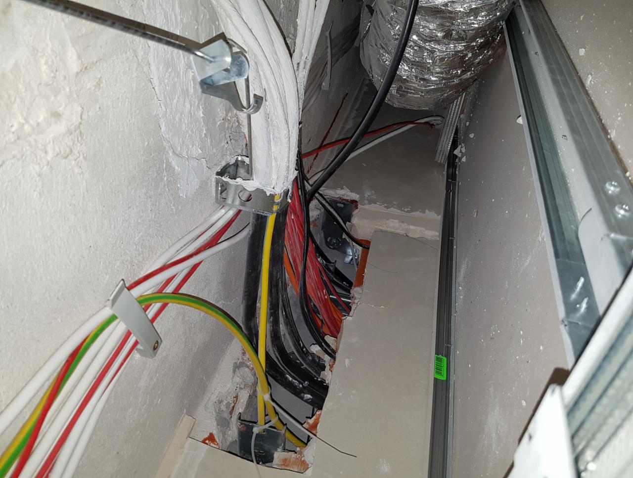 Instalacja elektryczna w trakcie remontu: widoczne kable w kolorowych izolacjach, metalowe uchwyty mocujące, fragment wentylacji z folii aluminiowej i profil konstrukcji ściany działowej.