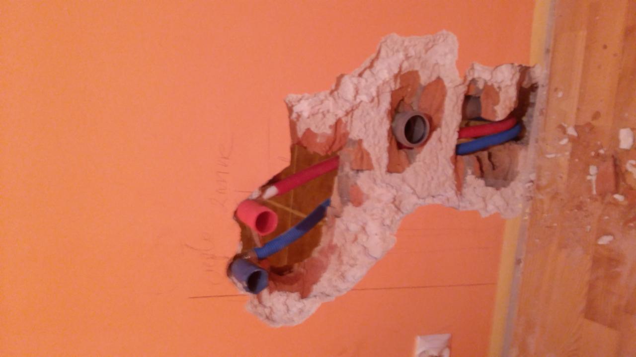 Odsłonięte rury instalacji wodnej (czerwona i niebieska) w wykuciu w pomarańczowej ścianie, widoczne ślady po wierceniu i resztki gruzu, fragment drewnianej podłogi po prawej stronie.