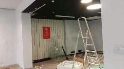 Wnętrze w trakcie prac wykończeniowych: surowe ściany, czarny sufit z oświetleniem punktowym, drabina aluminiowa, pionowe panele dekoracyjne na ścianie w głębi pomieszczenia, widoczne narzędzia...