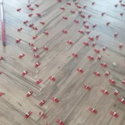 Układanie podłogi z płytek imitujących drewno w jodełkę, z użyciem czerwonych klipsów poziomujących, widoczna poziomica przy krawędzi.