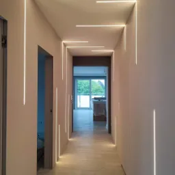 Nowoczesny korytarz z minimalistycznym oświetleniem LED wbudowanym w ściany i sufit, drewnopodobna podłoga, widok na jasne pomieszczenie w głębi.