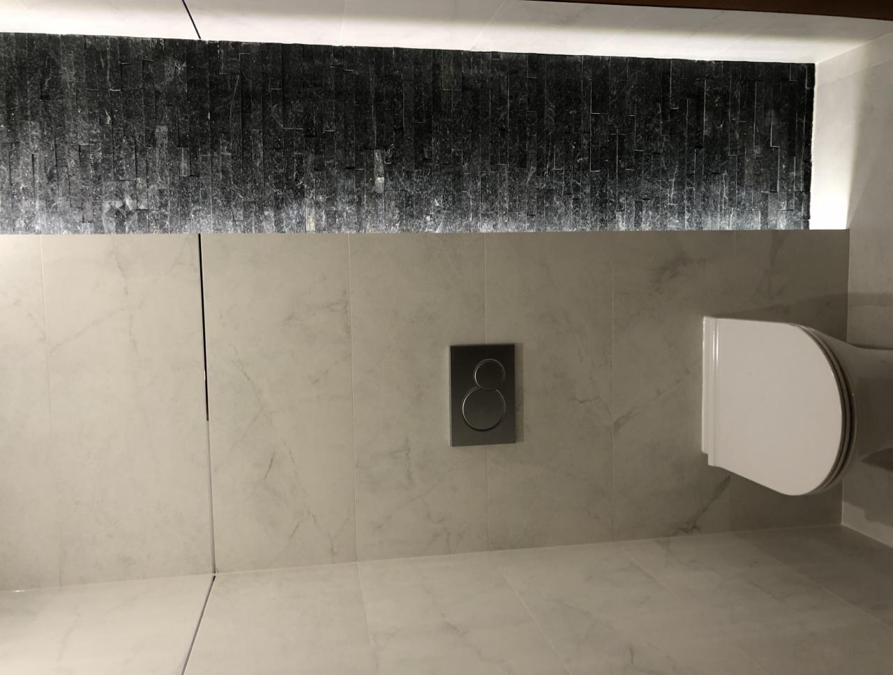 Nowoczesna toaleta podwieszana z szarą mozaiką kamienną podświetloną od góry oraz marmurowe płytki ścienne i podłogowe.