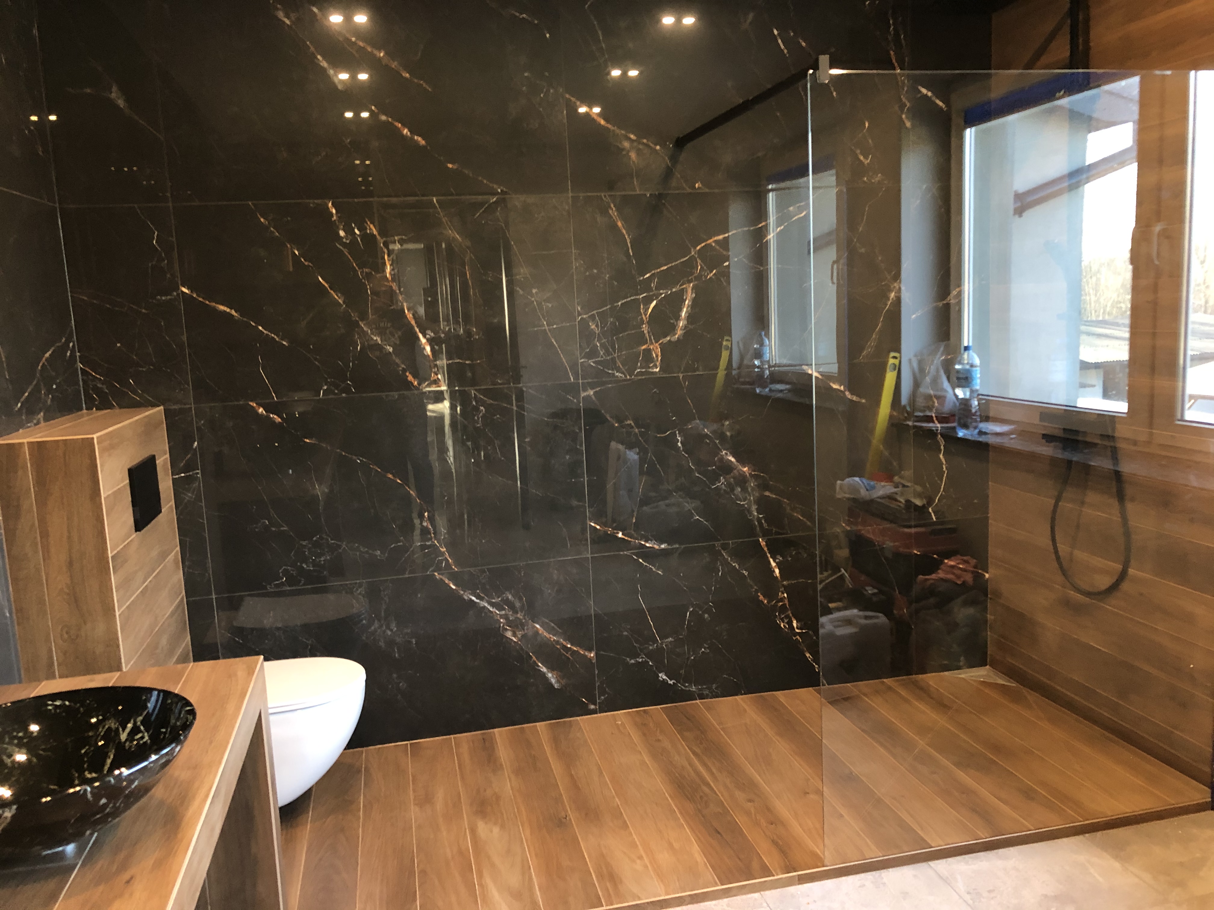 Wykończona łazienka z czarnymi płytkami imitującymi marmur z złotymi żyłami na ścianach, drewnopodobną podłogą, szklaną kabiną prysznicową i umywalką w stylu glamour.