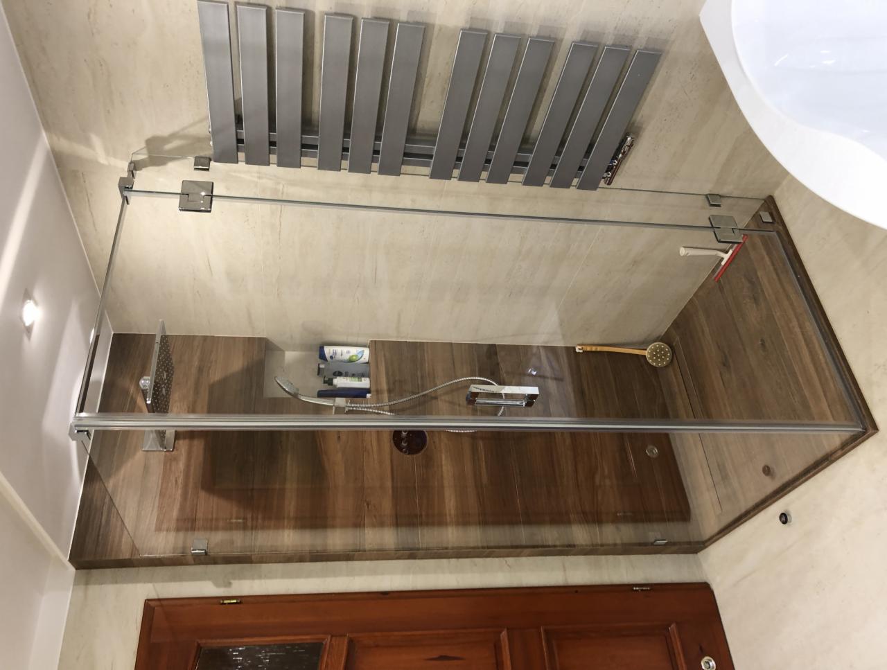 Szklana kabina prysznicowa z chromowanymi okuciami, drewnopodobne płytki na ścianie, grzejnik drabinkowy w łazience z widocznym fragmentem umywalki.