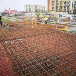 Siatka zbrojeniowa na deskowaniu, dwóch pracowników w odblaskowych kamizelkach i kaskach, budynki mieszkalne w tle.