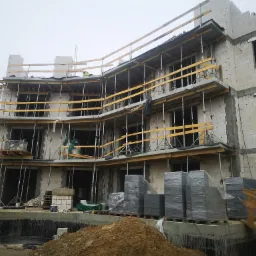 Wielopiętrowy budynek w trakcie budowy z białych bloczków, widoczne rusztowania, podpory stropowe i materiały budowlane na placu budowy w pochmurny dzień.