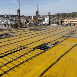 Mariusz Łapot budownictwo - Przygotowanie zbrojenia płyty fundamentowej na żółtym szalunku, widoczne pionowe pręty zbrojeniowe, w tle zakład kruszyw i pracownik w pomarańczowym ubraniu.