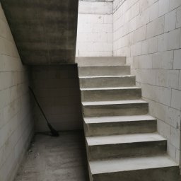 Mariusz Łapot budownictwo - Surowe betonowe schody wewnętrzne w trakcie budowy, widoczne ściany z białych bloczków, pod schodami oparta o ścianę miotła.