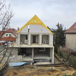 Mariusz Łapot budownictwo - Dwukondygnacyjny budynek w stanie surowym otwartym z żółtą konstrukcją dachu, widoczni dwaj pracownicy w żółtych kamizelkach, w tle zabudowa jednorodzinna i pochmurne niebo.