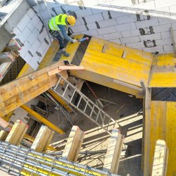 Mariusz Łapot budownictwo - Pracownik w żółtym kasku i kamizelce, stojący na żółtym szalunku, aplikuje uszczelniacz między bloczkami z betonu komórkowego w nowo powstającym budynku. Widoczna drabina, zbrojenie i betonowe...