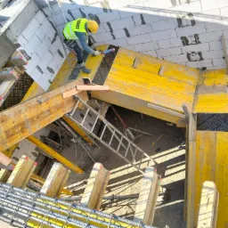 Pracownik w żółtym kasku i kamizelce, stojący na żółtym szalunku, aplikuje uszczelniacz między bloczkami z betonu komórkowego w nowo powstającym budynku. Widoczna drabina, zbrojenie i betonowe...