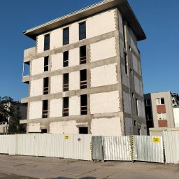 Mariusz Łapot budownictwo - Surowy budynek z betonu komórkowego w trakcie budowy, z widocznymi otworami okiennymi, otoczony metalowym ogrodzeniem na tle błękitnego nieba.