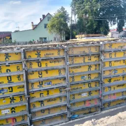 Szczegółowe ujęcie szalunków Doka, z widocznymi elementami mocującymi, na tle budynków i drzew, w słoneczny dzień. Widoczne zbrojenia.