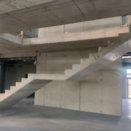 Surowe, betonowe schody w budynku w trakcie budowy. Widoczne ściany i sufit również z betonu, podkreślające minimalistyczny, industrialny charakter wnętrza.