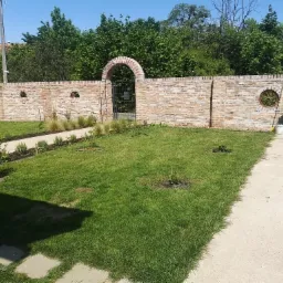 Długi, ceglany mur z łukowym przejściem i trzema półokrągłymi otworami, otaczający zielony trawnik z młodymi drzewkami i ścieżką z płyt betonowych, z widocznymi pojemnikami na śmieci po prawej...