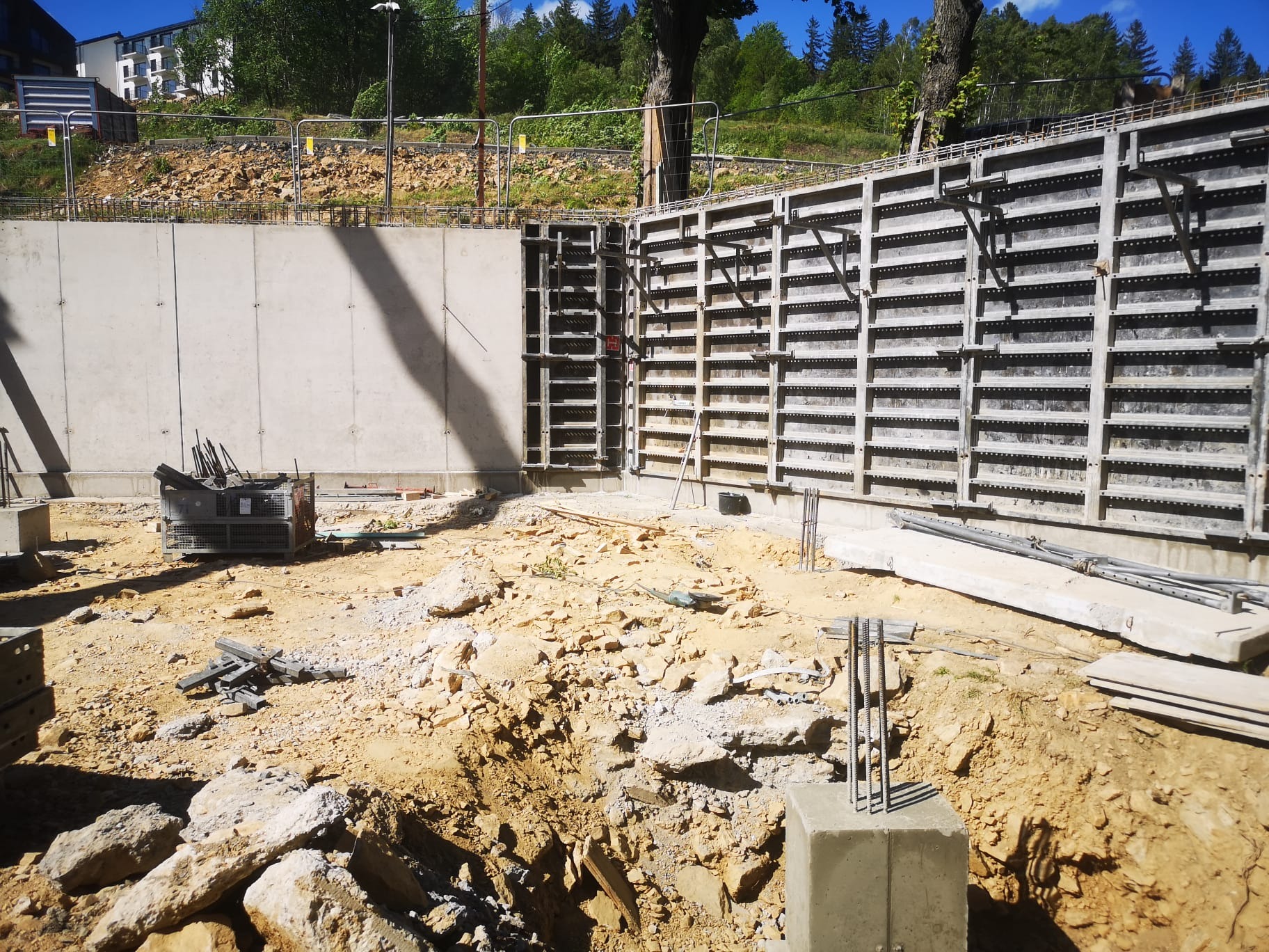 Szare betonowe ściany szalunkowe z widocznymi elementami zbrojenia na placu budowy w słoneczny dzień, z ziemią i gruzem w tle.