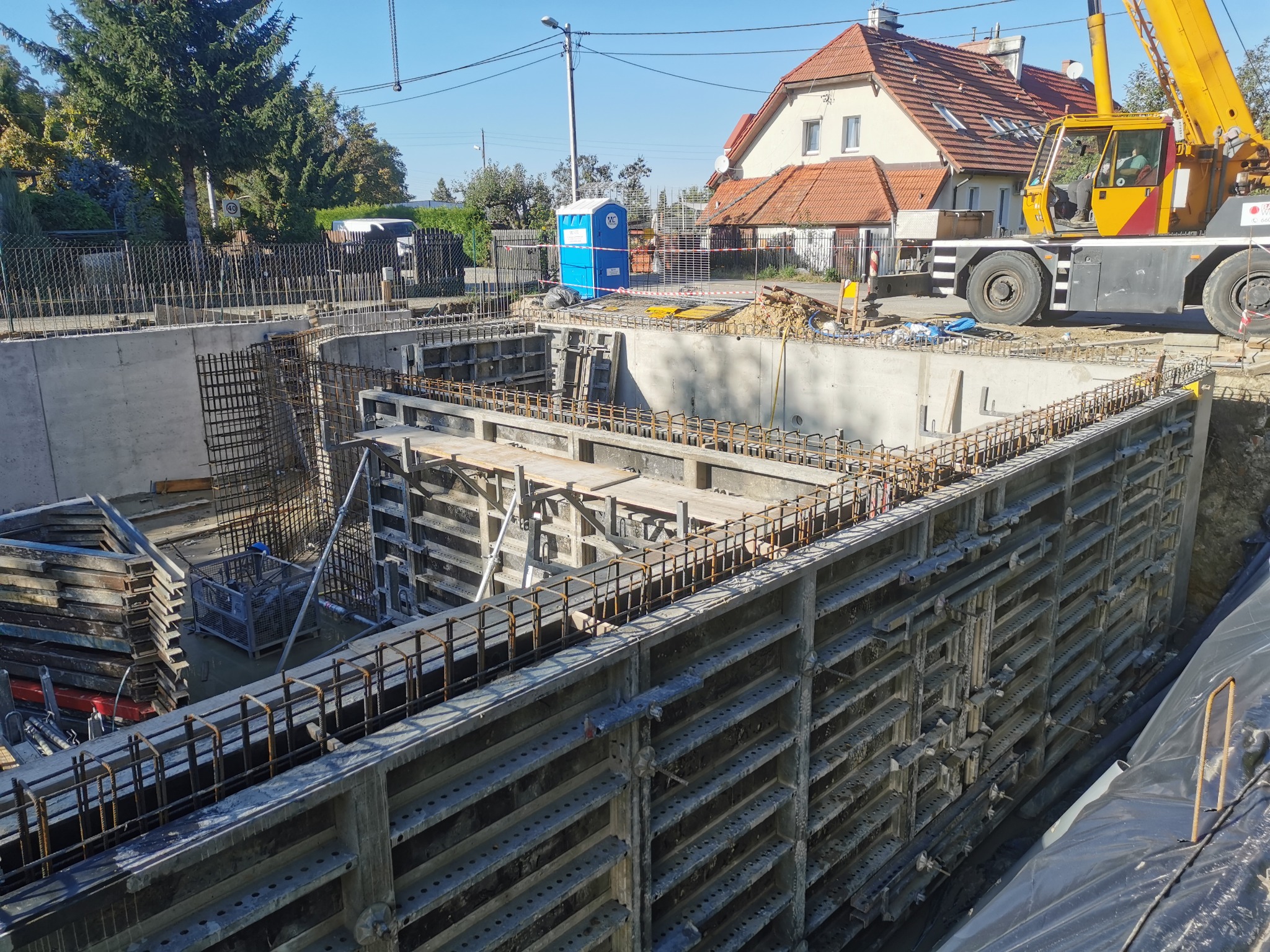 Wykop pod fundamenty z szalunkami i zbrojeniami, widoczny żuraw budowlany oraz przenośna toaleta w tle.