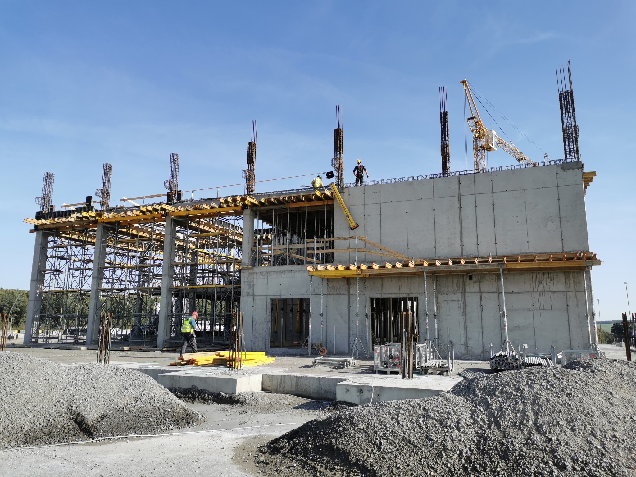 Budowa budynku żelbetowego w trakcie realizacji, widoczne szalunki, zbrojenia, betonowe ściany, żuraw budowlany i pracownicy w kaskach.