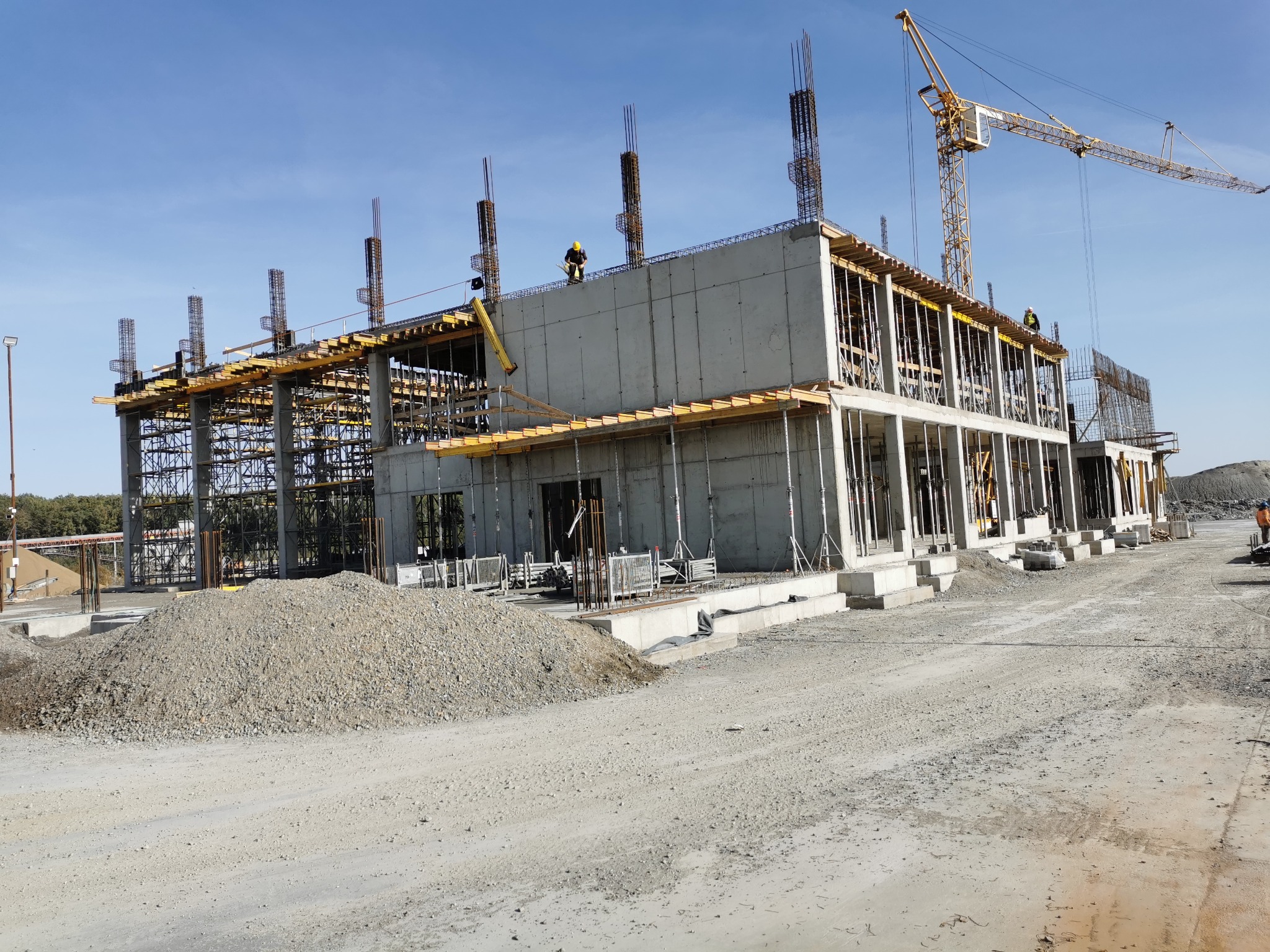 Wznoszenie dwukondygnacyjnego budynku z betonu, widoczne szalunki i zbrojenia, na budowie pracuje dźwig, a na górze budynku dwóch robotników w żółtych kaskach.