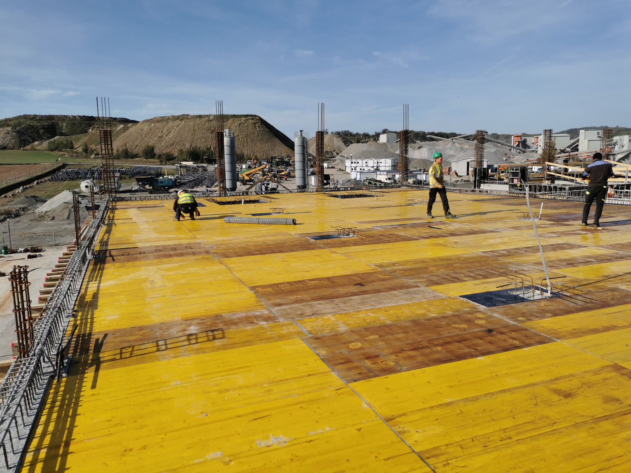 Widok z góry na plac budowy z żółtym szalunkiem i stalowymi zbrojeniami, pracownicy w odblaskowych kamizelkach i kaskach, w tle wzgórza i zabudowania przemysłowe.