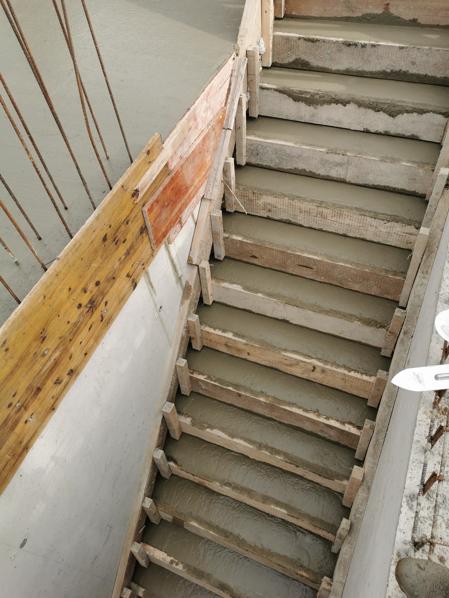 Wypełnione betonem szalunki drewniane schodów w trakcie budowy, widoczne wystające pręty zbrojeniowe i gładka powierzchnia betonu na ścianie obok.