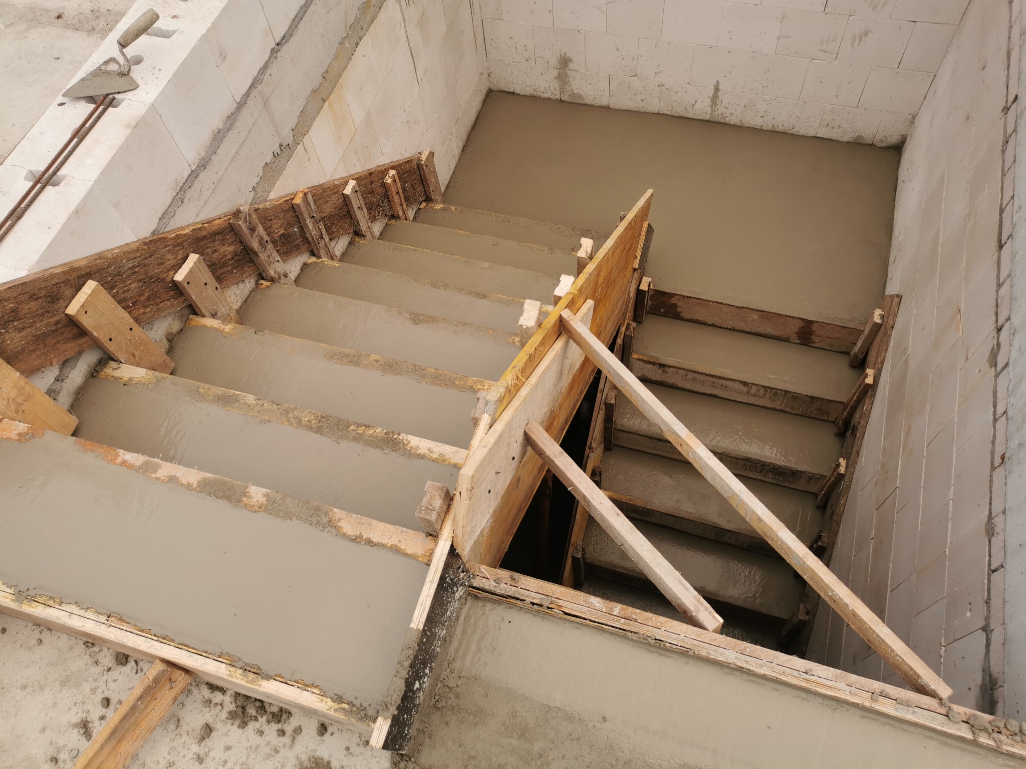 Wnętrze w trakcie budowy: zalane betonem schody ze szalunkiem drewnianym, widok z góry na mokry beton, ściany z bloczków betonowych, kielnia i pręty zbrojeniowe w tle.