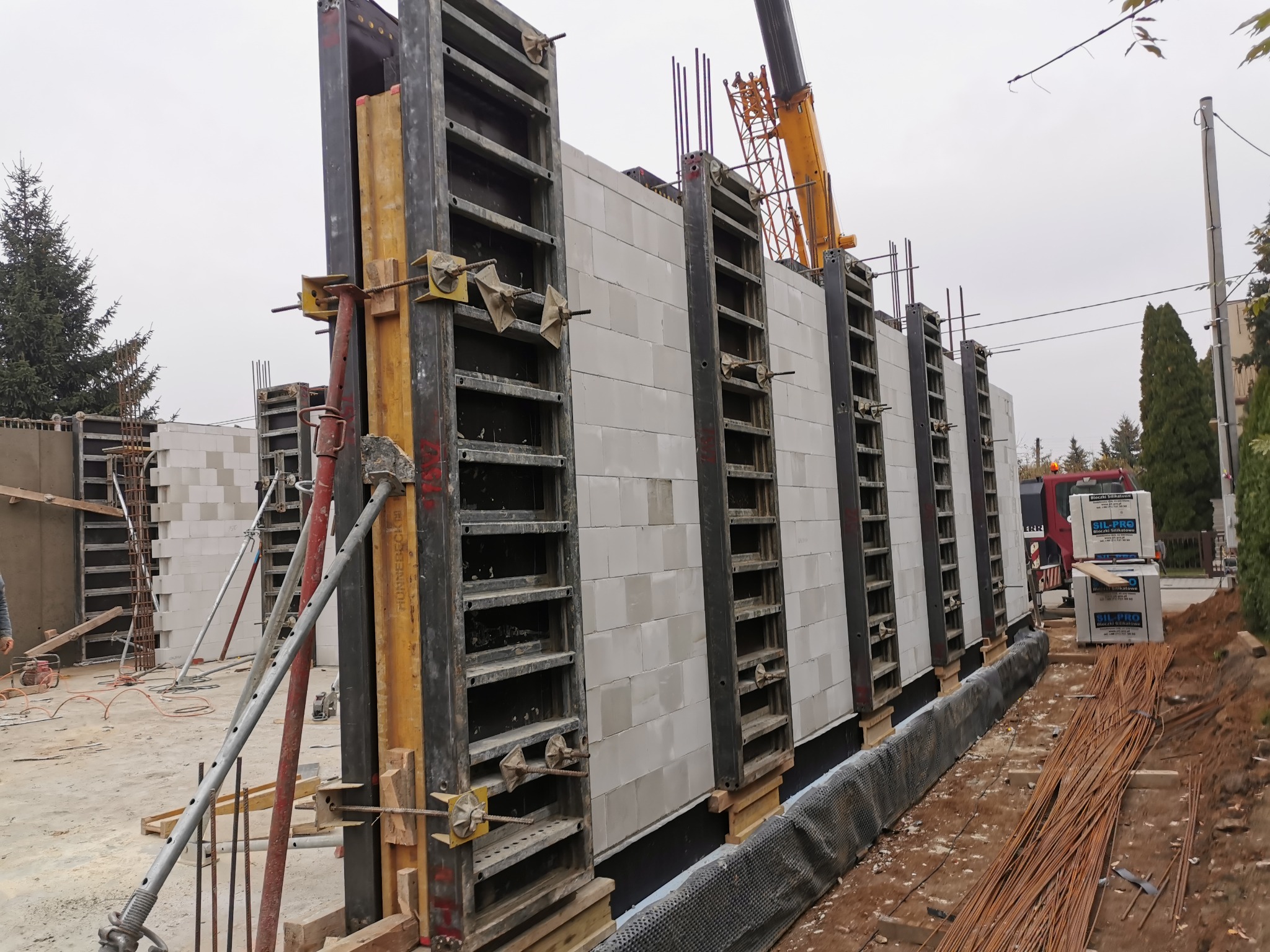 Wznoszenie ściany z białych bloczków silikatowych w szalunkach systemowych, widoczne zbrojenie pionowe i żuraw budowlany w tle.