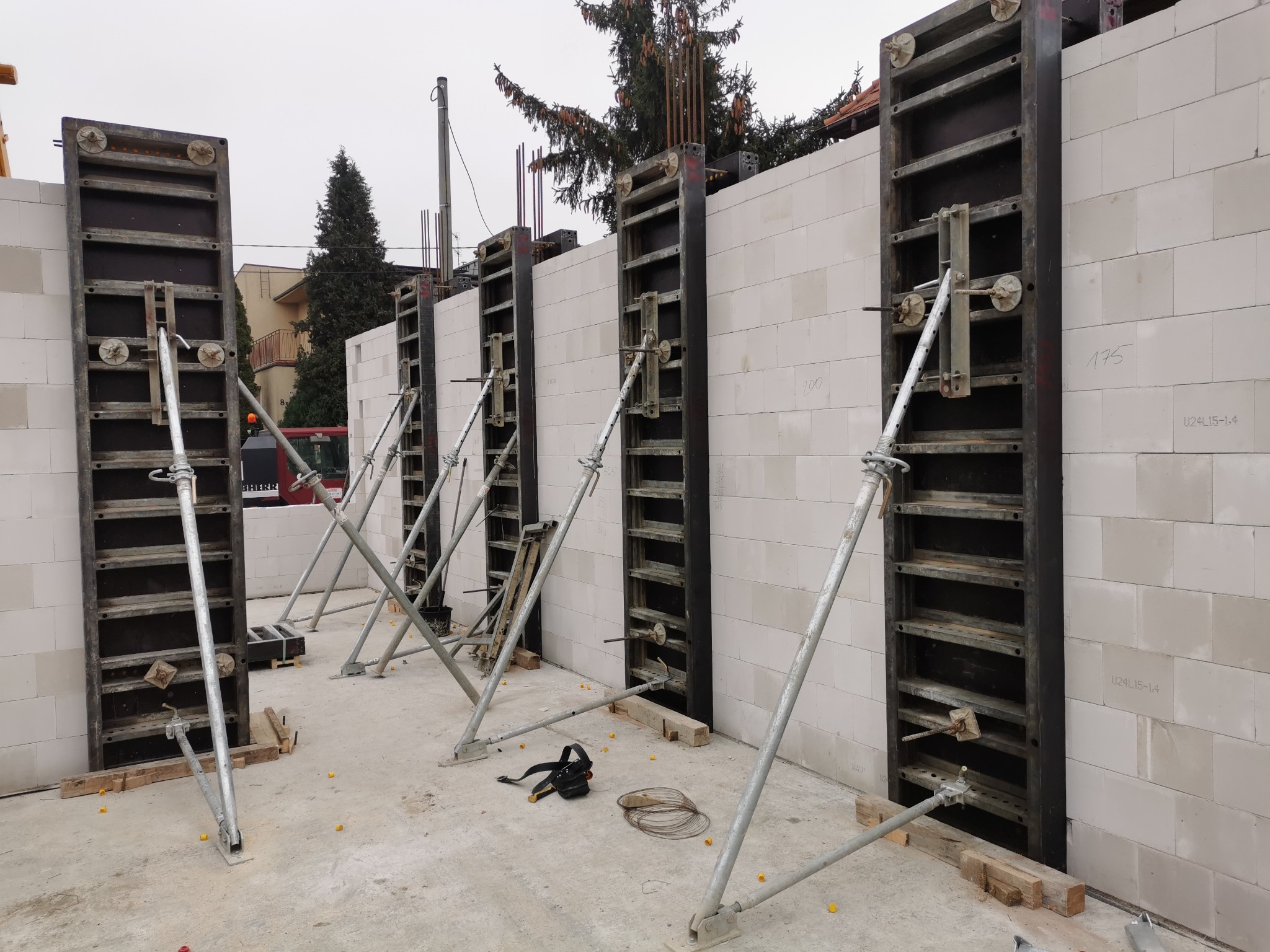 Wznoszenie ściany z bloczków, wzmocnionej szalunkami i metalowymi podporami, widok na plac budowy z elementami konstrukcyjnymi i narzędziami.