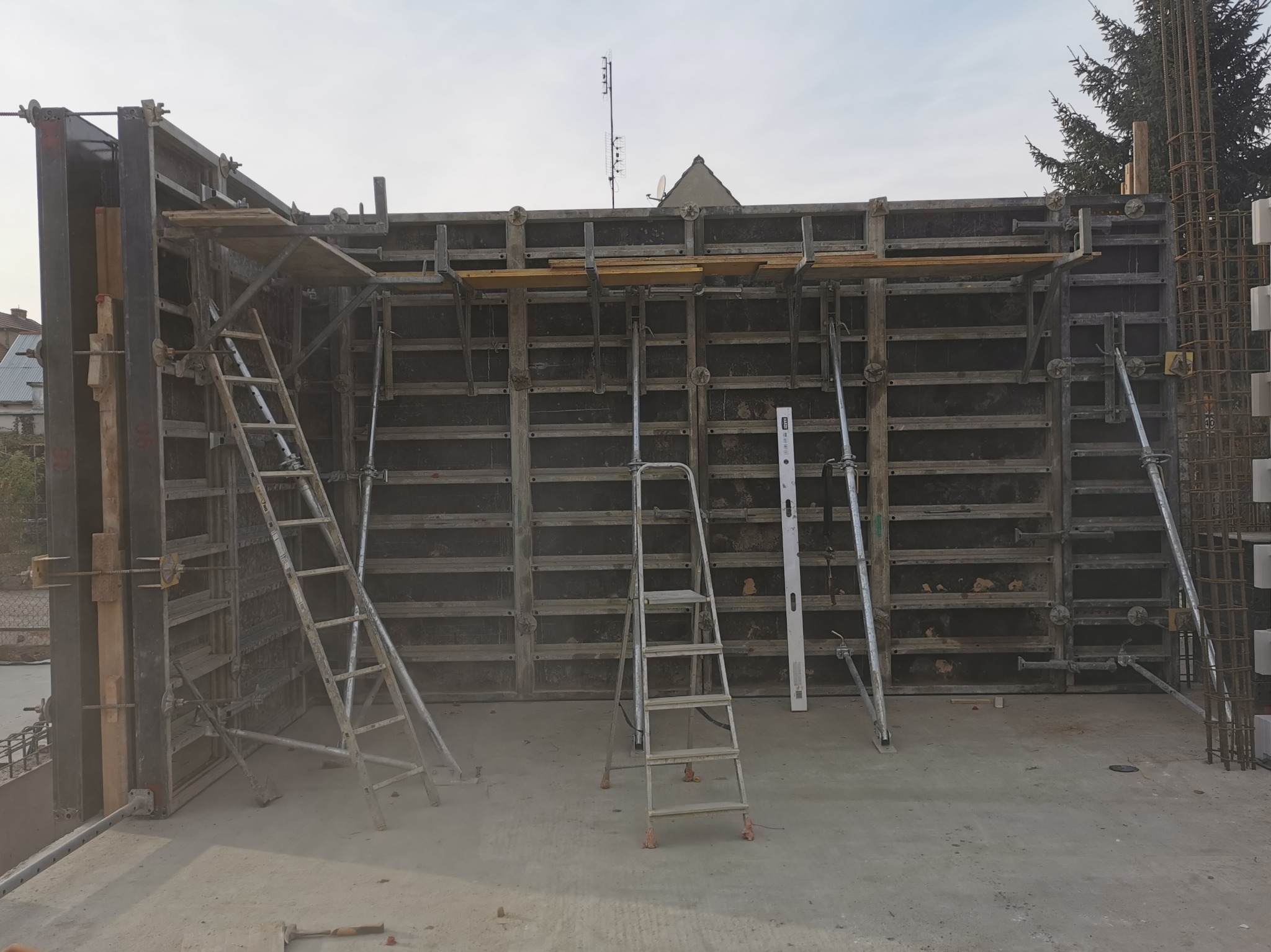 Konstrukcja szalunkowa z metalowymi elementami i drabinkami na placu budowy, widoczne zbrojenia i poziomica, w tle zabudowa mieszkalna.