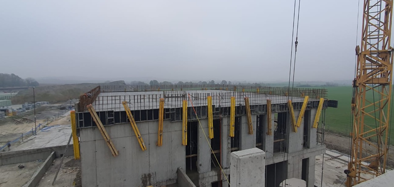 Wysoki budynek w trakcie budowy, z widocznym zbrojeniem na dachu i żółtymi podporami szalunkowymi przy ścianach, na tle pochmurnego nieba i krajobrazu.