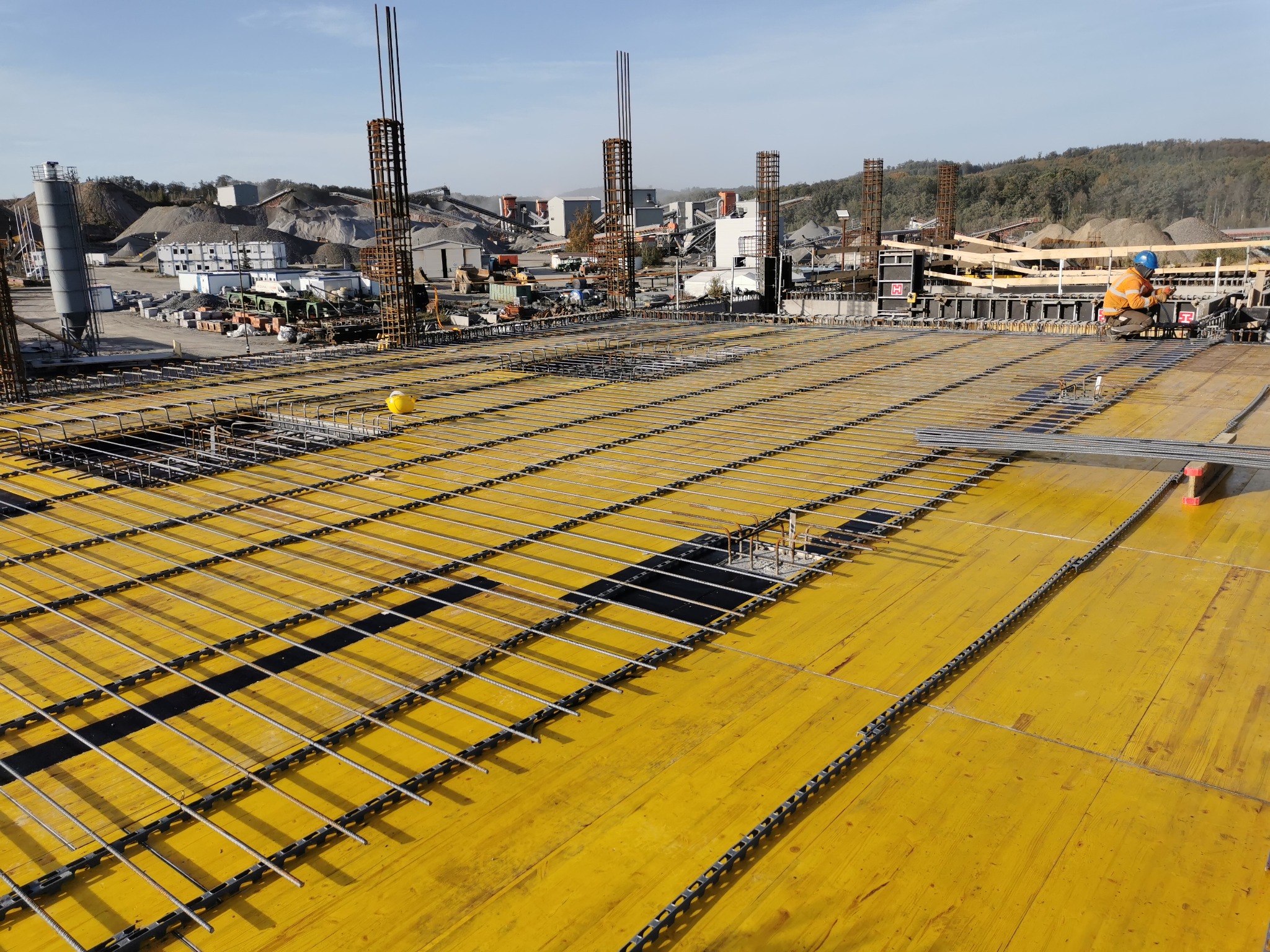 Przygotowanie zbrojenia płyty fundamentowej na żółtym szalunku, widoczne pionowe pręty zbrojeniowe, w tle zakład kruszyw i pracownik w pomarańczowym ubraniu.