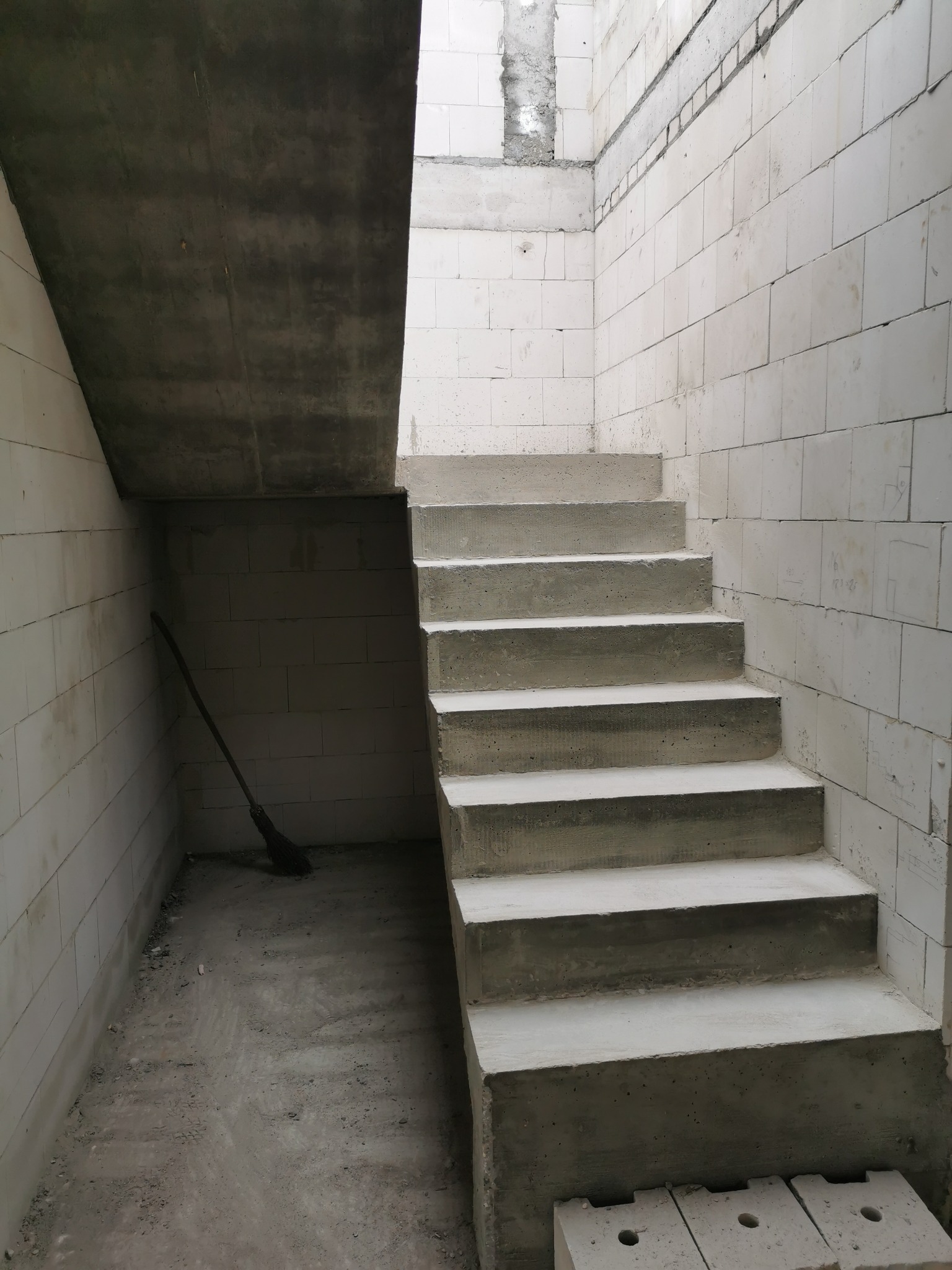 Surowe betonowe schody wewnętrzne w trakcie budowy, widoczne ściany z białych bloczków, pod schodami oparta o ścianę miotła.