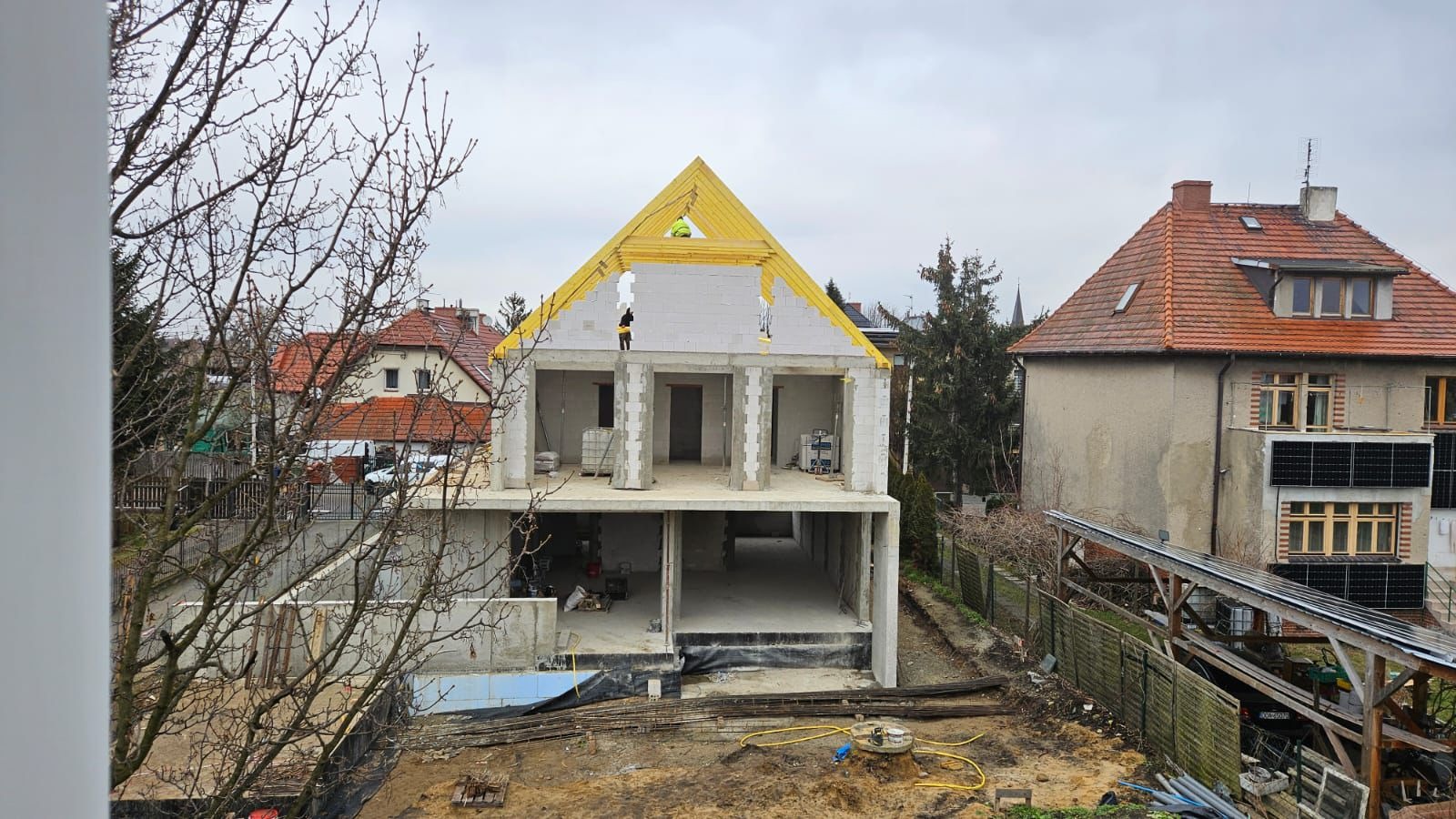 Dwukondygnacyjny budynek w stanie surowym otwartym z żółtą konstrukcją dachu, widoczni dwaj pracownicy w żółtych kamizelkach, w tle zabudowa jednorodzinna i pochmurne niebo.