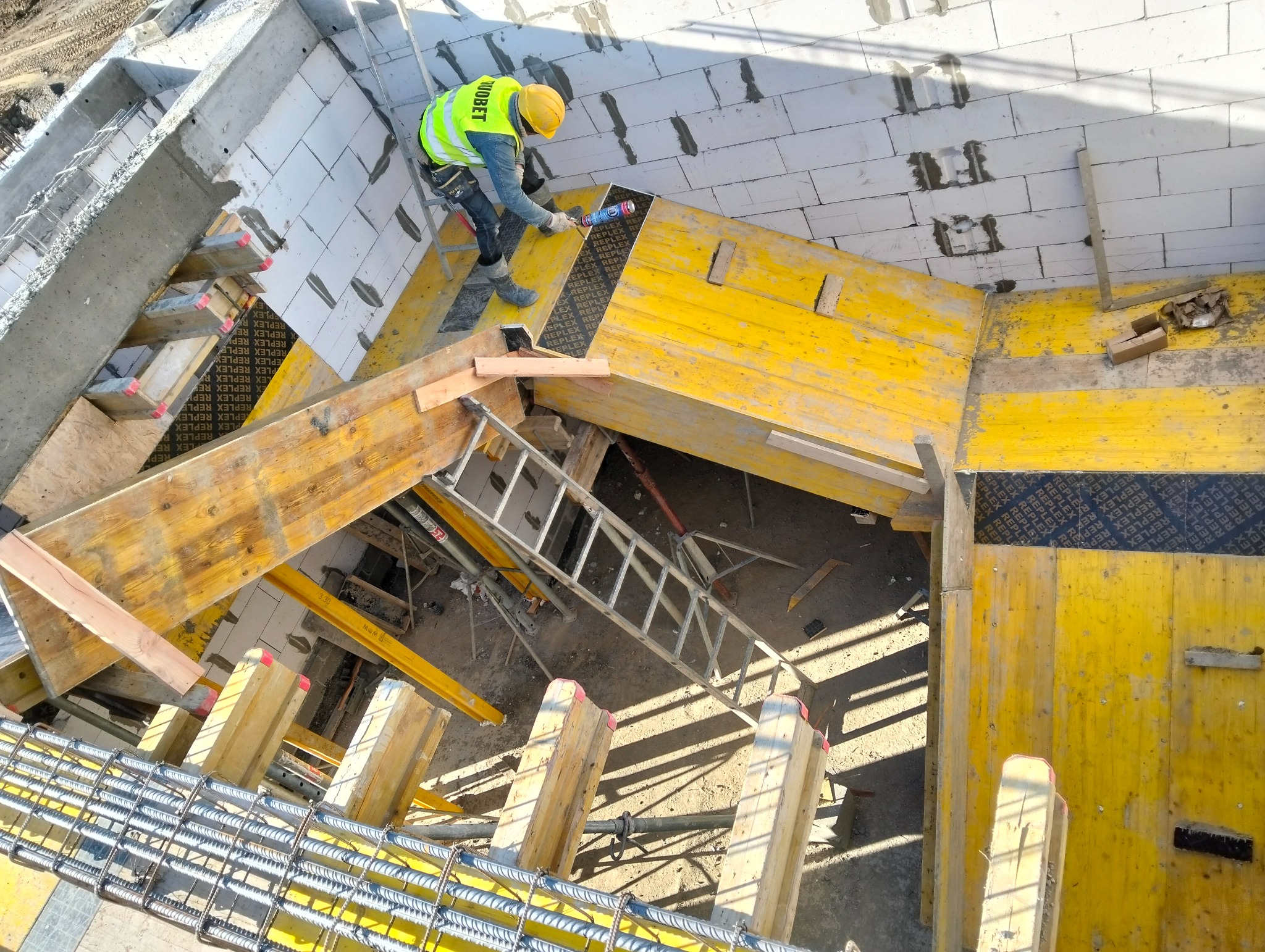 Pracownik w żółtym kasku i kamizelce, stojący na żółtym szalunku, aplikuje uszczelniacz między bloczkami z betonu komórkowego w nowo powstającym budynku. Widoczna drabina, zbrojenie i betonowe...