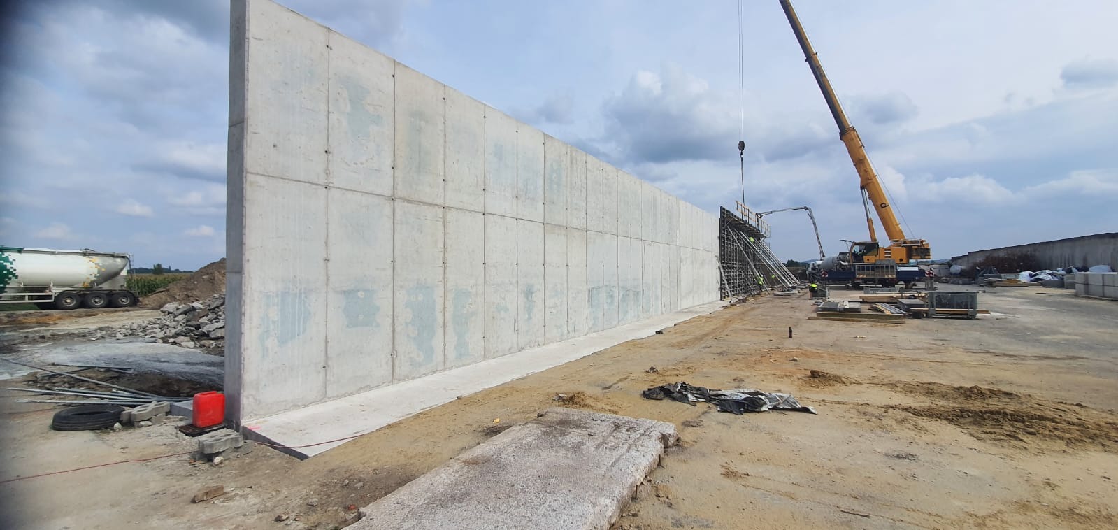 Wzniesiony mur oporowy z betonu na placu budowy, widoczny żuraw i betonowóz. Konstrukcja składa się z prefabrykowanych elementów, widoczne ślady łączeń.