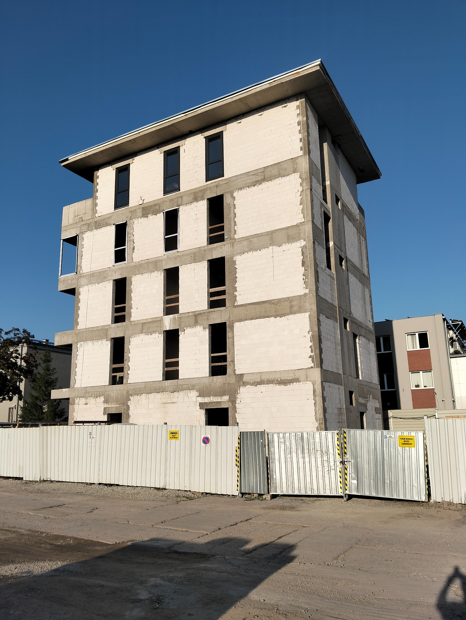 Surowy budynek z betonu komórkowego w trakcie budowy, z widocznymi otworami okiennymi, otoczony metalowym ogrodzeniem na tle błękitnego nieba.