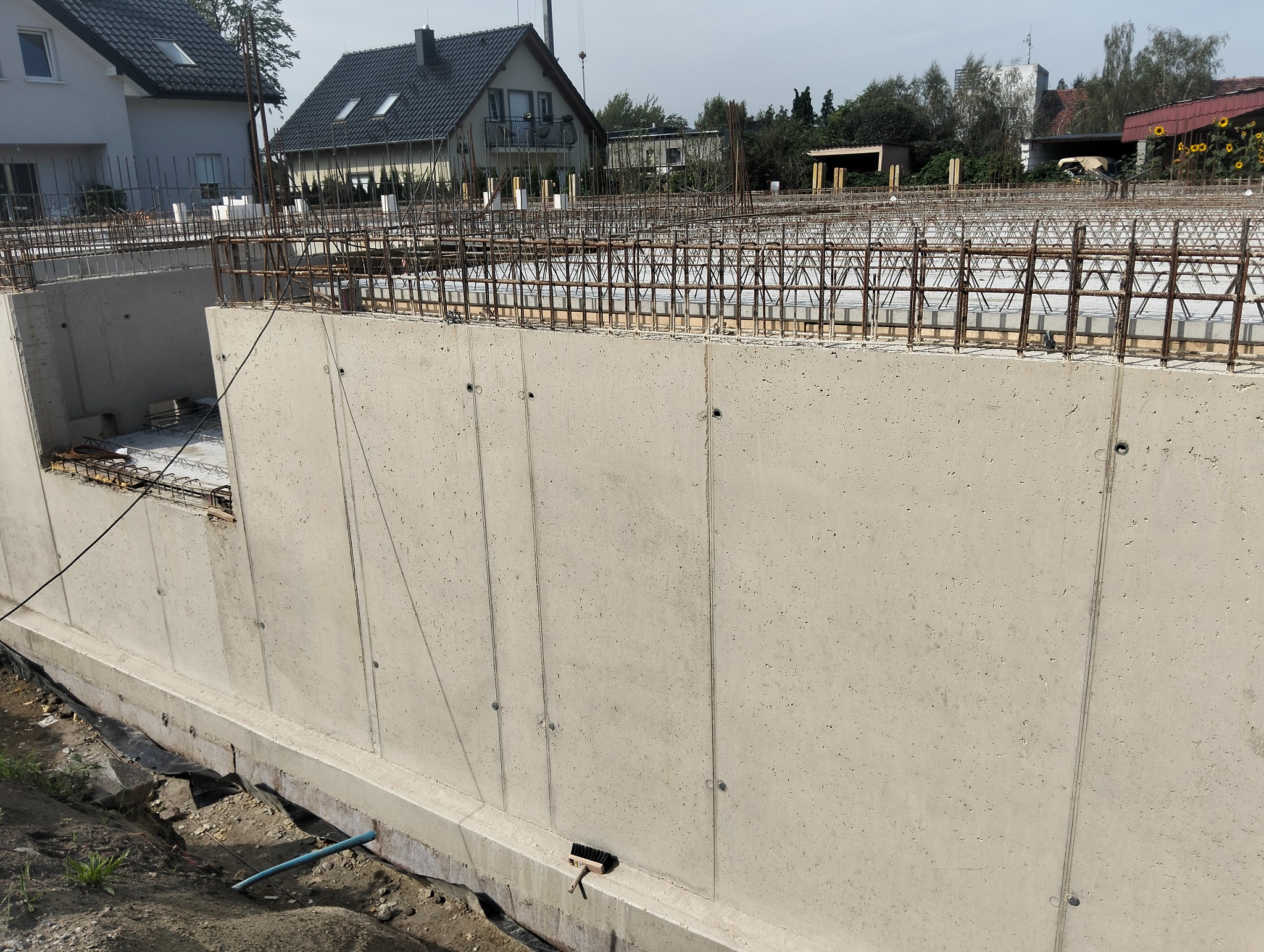 Świeżo wylany, szalowany fundament z widocznym zbrojeniem na tle domów jednorodzinnych i zieleni. Elementy szalunku i poziomowania widoczne w tle.