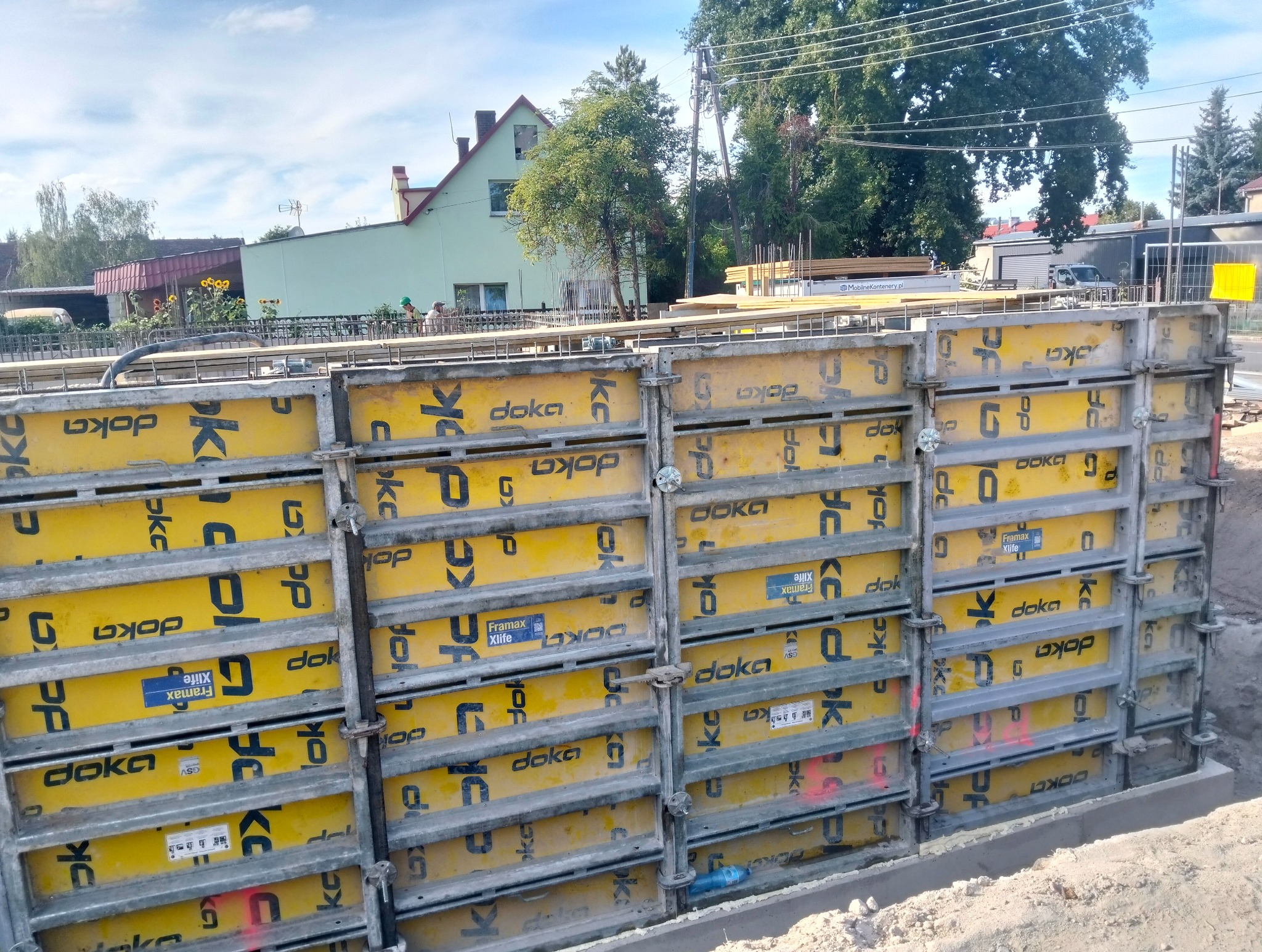 Szczegółowe ujęcie szalunków Doka, z widocznymi elementami mocującymi, na tle budynków i drzew, w słoneczny dzień. Widoczne zbrojenia.