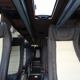 Wnętrze autobusu z brązowo-kremowymi skórzanymi fotelami, widok na schodki i okno dachowe.