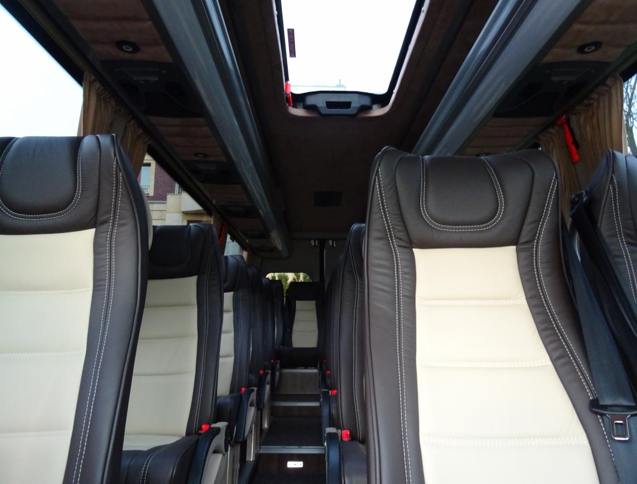 Wnętrze autobusu z brązowo-kremowymi skórzanymi fotelami, widok na schodki i okno dachowe.