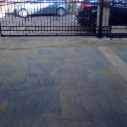 Ułożona kostka brukowa na podjeździe, posypana piaskiem, z bramą w tle i widocznymi fragmentami zaparkowanych samochodów.