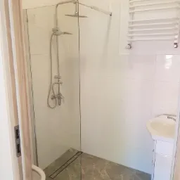 Nowoczesna kabina prysznicowa walk-in z deszczownicą i odpływem liniowym w wykończonej łazience z białymi płytkami i szarą marmurową podłogą, widok zza lekko uchylonych drewnianych drzwi.