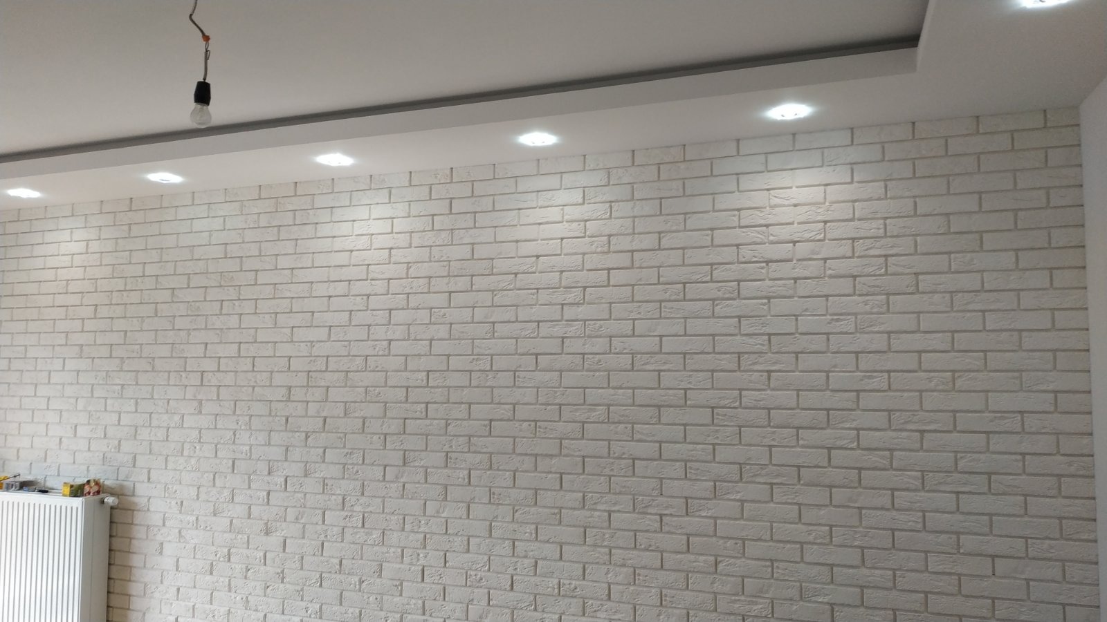 Ściana wykończona białą cegłą dekoracyjną podświetlona punktowymi lampami sufitowymi, widoczny fragment grzejnika i wisząca żarówka.
